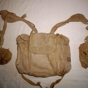 Op de afbeelding: Een beige canvas militaire veldrugzak met een centraal rechthoekig compartiment met klepsluiting, twee kleinere zijvakken en verstelbare schouderbanden met metalen gespen. De stof vertoont duidelijke tekenen van slijtage en verkleuring.