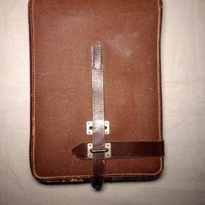 May include: A rectangular brown leather case with a textured surface and rounded corners. The case features a vertical leather strap secured by a horizontal leather band and silver-toned metal hardware, including rivets and a buckle-style closure.