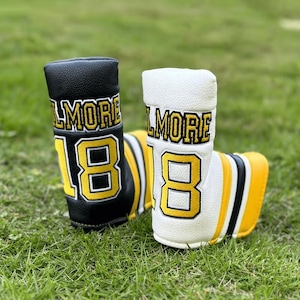 May include: Two golf club head covers, one black and one white, with the word "LMORE" and the number "18" in yellow. The covers have a football jersey design with yellow and black stripes.