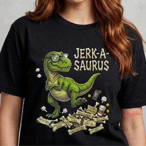 Puede incluir: Camiseta negra con un dinosaurio verde con gafas y el texto "JERK-A-SAURUS". El diseño incluye huesos con frases como "Ghosting" y "Red Flag". Una camiseta gráfica humorística.