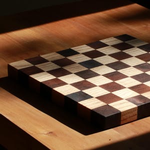 May include: A square wooden chessboard featuring a classic alternating checkerboard pattern. The board is constructed from light-colored wood squares and dark-colored wood squares, creating a high-contrast grid design with visible wood grain textures.