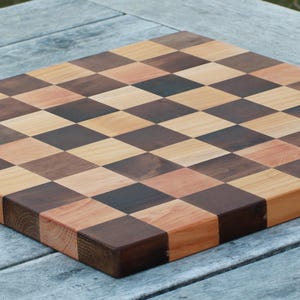 Könnte beinhalten: Ein quadratisches Holzschneidebrett mit einem Schachbrettmuster aus abwechselnd hellbraunen und dunkelbraunen Holzblöcken. Das Brett hat eine glatte, flache Oberfläche mit sichtbarer Holzmaserung und geraden Kanten in einem Gitterdesign.