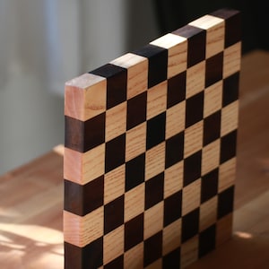 May include: A square wooden cutting board featuring a checkerboard pattern of alternating light beige and dark brown wood squares. The board is positioned vertically, displaying the grid design on its front face and its thick, layered side edge.