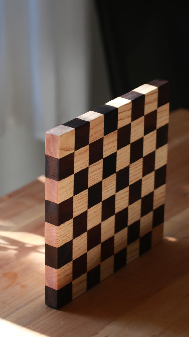 May include: A square wooden cutting board featuring a checkerboard pattern of alternating light beige and dark brown wood squares. The board is positioned vertically, displaying the grid design on its front face and its thick, layered side edge.