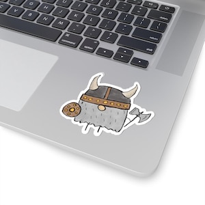 May include: A sticker featuring a cartoon Viking character with a horned helmet, shield, and axe. The Viking has a gray beard and is wearing a helmet with gold accents. The shield is brown with gold details. The sticker is on a laptop.
