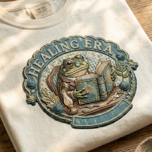 May include: Cream-colored t-shirt featuring an embroidered design of a frog reading a book, with the text "HEALING ERA" in a circular frame. The frog is wearing a jacket and the book is open. The shirt has a tag that says "COMFORT COLORS".