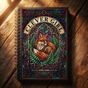 May include: A spiral-bound notebook with a stained-glass style cover. The cover features a fox illustration surrounded by green leaves, with the words "CLEVER GIRL" arched above. The color palette includes orange, green, purple, and gold.
