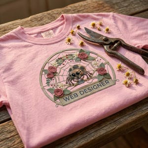Può includere: Una maglietta rosa con un design ricamato dettagliato con un ragno in una ragnatela, circondato da fiori. Il testo "WEB DESIGNER" è sotto. Forbici di metallo vintage e piccoli fiori sono accanto alla maglietta.