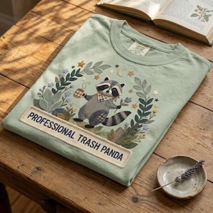 Könnte beinhalten: Hellgrünes T-Shirt mit einer Stickerei eines Waschbären, der eine Kaffeetasse und eine Blume hält, umgeben von Blättern, Sternen und Monden. Der Text "PROFESSIONAL TRASH PANDA" ist auf einem rechteckigen Aufnäher zu sehen.