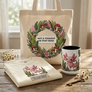 May include: A cream-colored tote bag, t-shirt, and mug set featuring a pink axolotl design. The tote bag has a wreath design with the text "NOT A THOUGHT IN THAT HEAD". The t-shirt and mug also have the axolotl and text.