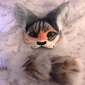Puede incluir: Una máscara de gato con una cola a juego. La máscara presenta un diseño pintado con pelaje naranja, gris y blanco. La cola es esponjosa con una mezcla de blanco, marrón y gris. Se ve un pequeño cierre plateado.