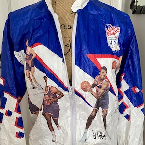 May include: A vintage, full-zip jacket in white, blue, and red, featuring basketball player graphics and signatures. The jacket has a white collar and a USA Olympic logo. The design includes diagonal stripes and geometric patterns.