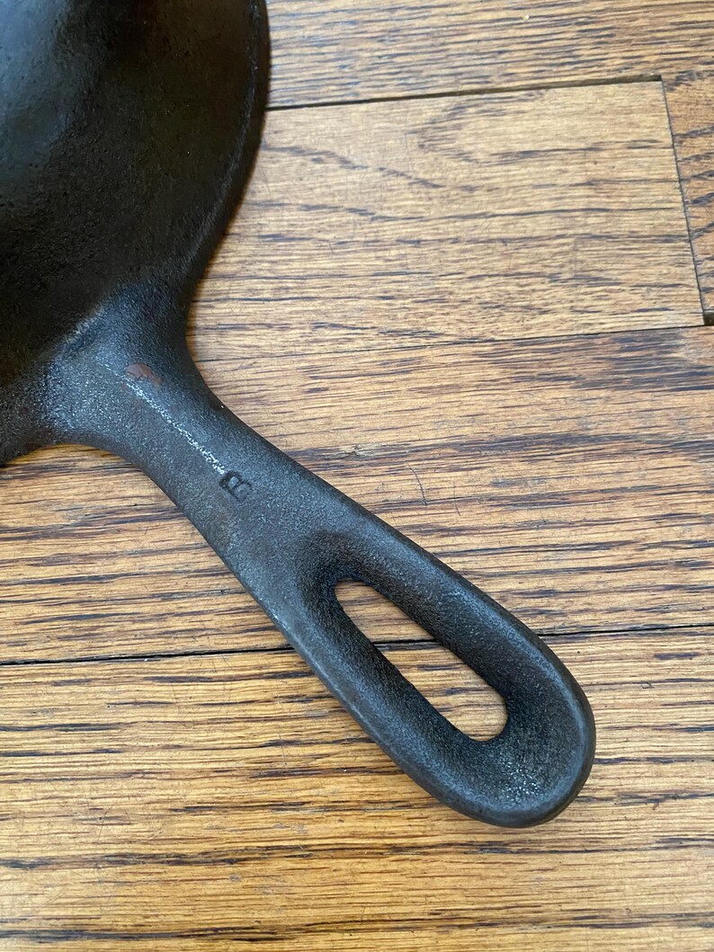May include: A black cast iron skillet with a single handle. The handle has a small hole in it. The skillet is on a wooden surface.