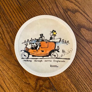 May include: Round ceramic coaster with a vintage illustration of a car driving through a town. The car is orange, and the illustration has a cream background with the text "walking through merrie Englande."
