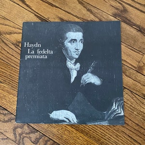 May include: A square, grayscale album cover featuring a portrait of a man in a dark suit and white cravat. The text "Haydn La fedelta premiata" is printed in white on the left side of the cover. The album is resting on a wooden surface.