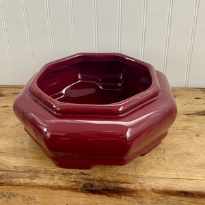 May include: A burgundy-colored, octagonal ceramic planter with a glossy finish. The planter has a wide, open top and a stepped base. It sits on a wooden surface.