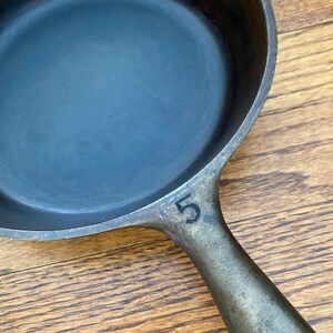 May include: A cast iron skillet with a black handle and a number "5" stamped on the side. The skillet is on a wooden surface.