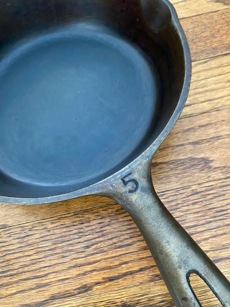 May include: A cast iron skillet with a black handle and a number "5" stamped on the side. The skillet is on a wooden surface.