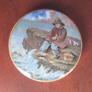 May include: A round ceramic tile with a painted scene of a person sitting on a rocky cliff by the ocean. The person is wearing a brown coat and a hat. The ocean is blue and white with waves crashing against the rocks. There is a basket and a few fish on the rocks.