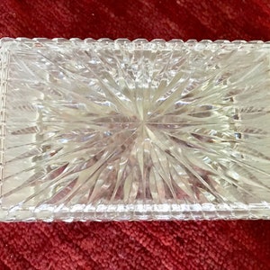 May include: A clear glass box with a cut-glass design. The box has a square shape and a raised edge.