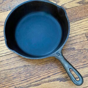 May include: A black cast iron skillet with a 5 stamped on the handle. The skillet is sitting on a wooden surface.