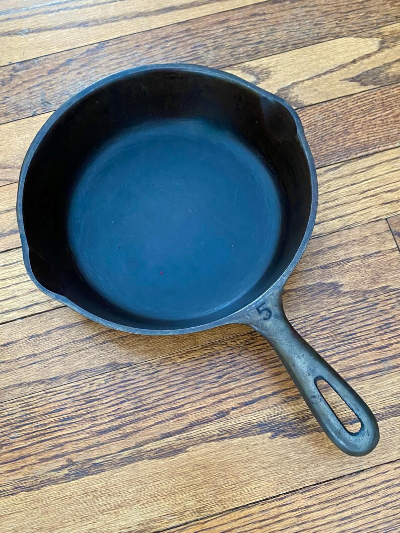 May include: A black cast iron skillet with a 5 stamped on the handle. The skillet is sitting on a wooden surface.