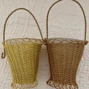 May include: Two decorative wire baskets with handles. One basket is gold, the other is a darker bronze color. Both have a woven design and a clear glass insert. The handles are made of twisted wire.