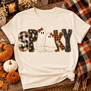 May include: A cream-colored short-sleeve t-shirt featuring the word SPOOKY printed across the chest. The letters are designed with various patterns including black and white buffalo plaid, black floral, and orange plaid, with two white ghost figures.