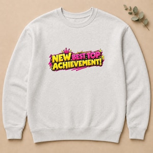 Puede incluir: Una sudadera de cuello redondo gris claro con un estampado gráfico en el pecho. El diseño muestra el texto "NEW BEST TOP ACHIEVEMENT!" en letras llamativas de estilo cómic en amarillo y rosa, con una pequeña corona rosa y estrellas.
