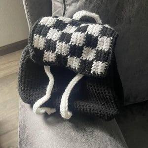May include: A handmade crochet backpack in black and white. The top flap features a checkerboard pattern, while the main body is solid black. White drawstrings and a handle complete the design. The backpack is sitting on a gray surface.