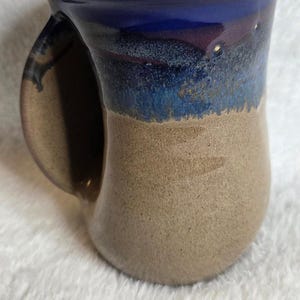 Puede incluir: Una taza de cerámica hecha a mano con un diseño único. La taza presenta un borde azul oscuro que se desvanece en un medio moteado azul y marrón, y una base marrón. La taza tiene un asa curva.