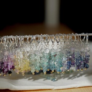 May include: A collection of colorful gemstone earrings displayed on a metal rod. The earrings feature a variety of colors, including purple, yellow, green, and blue, with small gold-colored beads at the bottom. The earrings are hanging on a white surface.