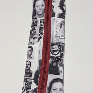 May include: A rectangular pencil case with a burgundy zipper and a black and white print featuring mugshots and newspaper clippings. The text "THI Face of Blind T" is visible on the case.