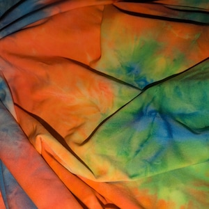 May include: A close-up of a tie-dye fabric with vibrant colors. The fabric features a blend of orange, green, blue, and yellow hues, creating a swirling, abstract pattern. The fabric is folded and draped, showing the texture and flow of the material.