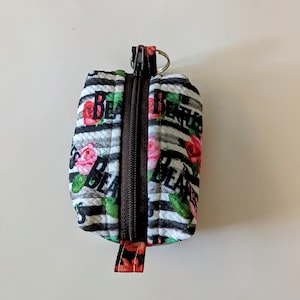May include: A small, rectangular zippered pouch with a black and white striped pattern. The pouch features pink roses and the word "BEATLES" in black. A brown zipper runs down the center, and a small metal ring is attached at the top.