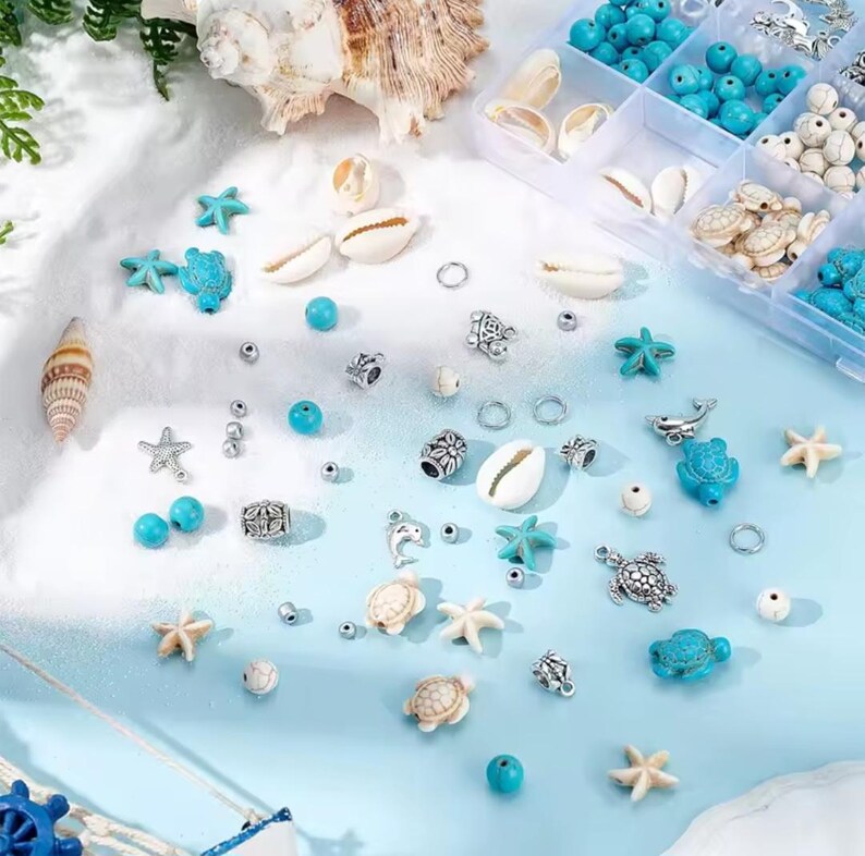 May include: A collection of jewelry making supplies including turquoise and white beads, silver-toned metal charms shaped like sea turtles, dolphins, and starfish, and small white seashells scattered on a light blue and white surface.