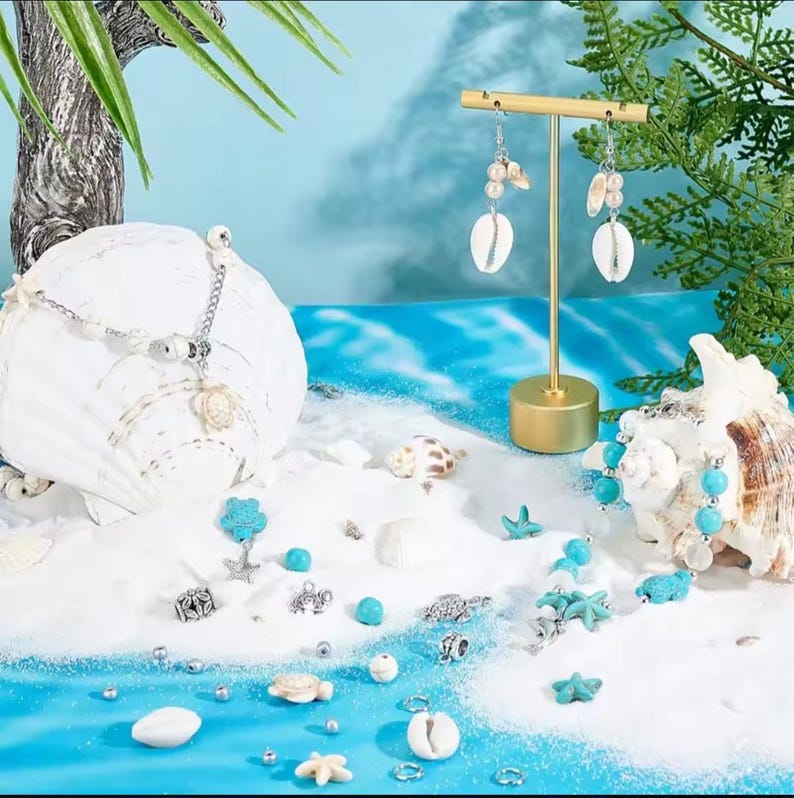 May include: A collection of jewelry featuring a silver-toned necklace with a turtle pendant, a pair of drop earrings with white shells and pearls, and various loose turquoise beads, silver charms, and small shells arranged on white sand.