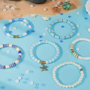 May include: A collection of five bracelets featuring flat circular clay beads in shades of blue and white, interspersed with gold-toned metallic spacers. One bracelet includes a blue starfish charm, and another consists of white pearl-like beads.