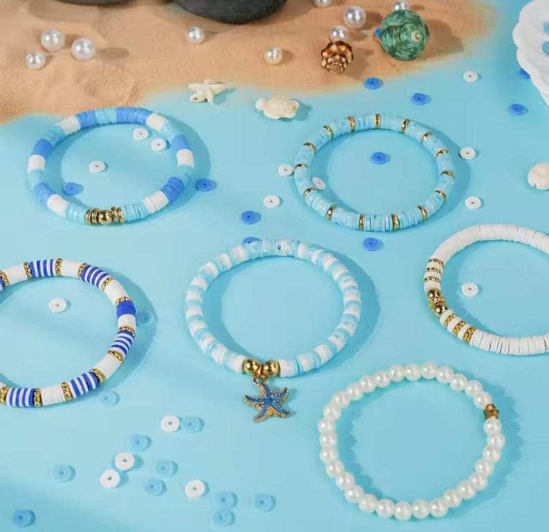 May include: A collection of five bracelets featuring flat circular clay beads in shades of blue and white, interspersed with gold-toned metallic spacers. One bracelet includes a blue starfish charm, and another consists of white pearl-like beads.