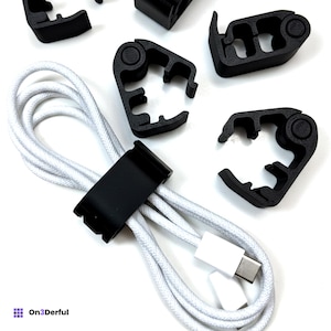May include: A collection of black plastic cable management clips with a textured finish, featuring a hinged design and circular pivot points. One clip is shown securing a white braided charging cable with a USB-C connector, while others are loose.