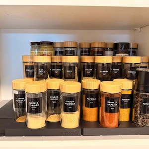 May include: A collection of spice jars with wooden lids, neatly arranged on black shelves inside a white cabinet. The jars are labeled with various spice names, including garlic powder, paprika, and pepper. The overall theme is kitchen organization.