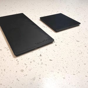 Peut inclure: Deux accessoires de cuisine rectangulaires noirs, l'un plus grand que l'autre, sont présentés sur une surface blanche mouchetée. Le plus grand article porte le mot "Spicefinity" en relief.