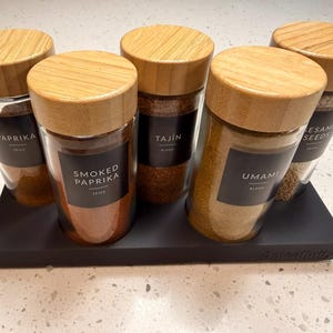 Peut inclure: Ensemble de cinq pots à épices en verre avec couvercles en bambou, chacun rempli d'épices différentes et étiqueté. Les pots sont disposés sur un plateau noir. Épices : paprika, paprika fumé, Tajin, mélange umami et graines de sésame.