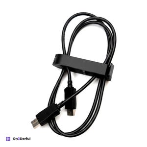 Puede incluir: Un cable USB negro enrollado y asegurado con un clip de gestión de cables de plástico negro. El cable cuenta con dos conectores USB rectangulares en los extremos. El texto On3Derful es visible en la esquina inferior izquierda.