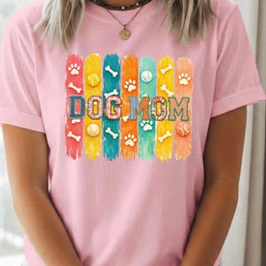 Colorful dog mom graphic with vertical painted stripes, paw prints, bones, and tennis balls in a playful patchwork style design for shirts and sublimation.