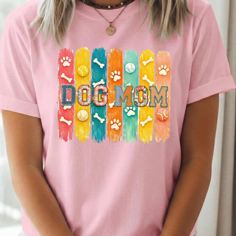 Colorful dog mom graphic with vertical painted stripes, paw prints, bones, and tennis balls in a playful patchwork style design for shirts and sublimation.