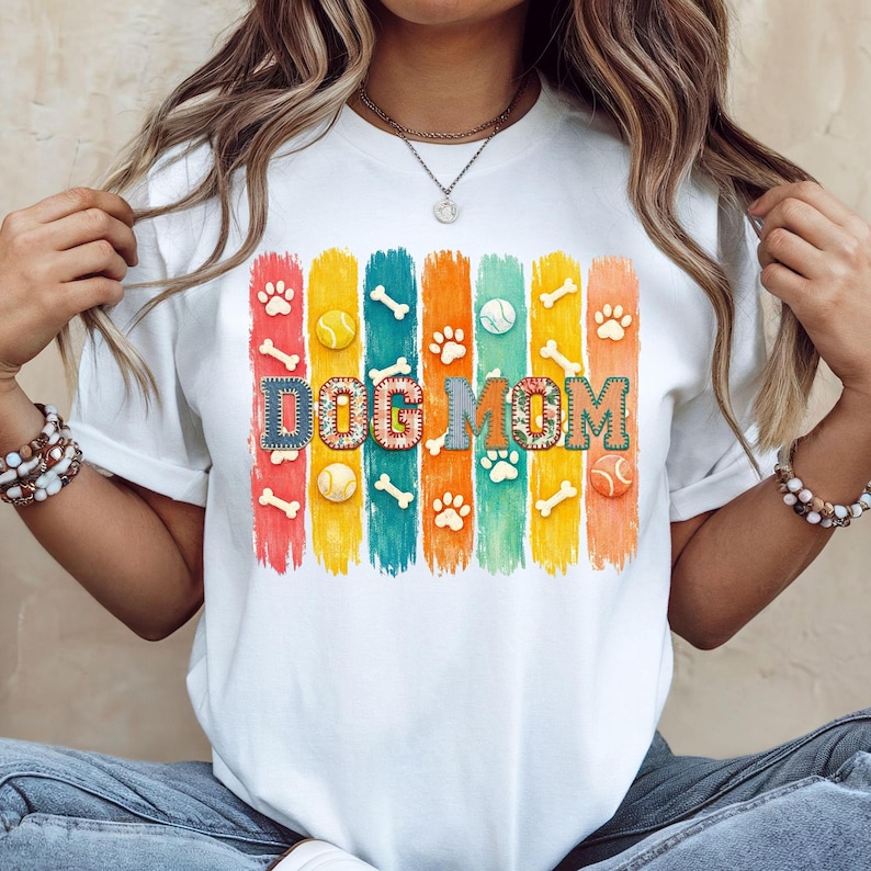 Colorful dog mom graphic with vertical painted stripes, paw prints, bones, and tennis balls in a playful patchwork style design for shirts and sublimation.