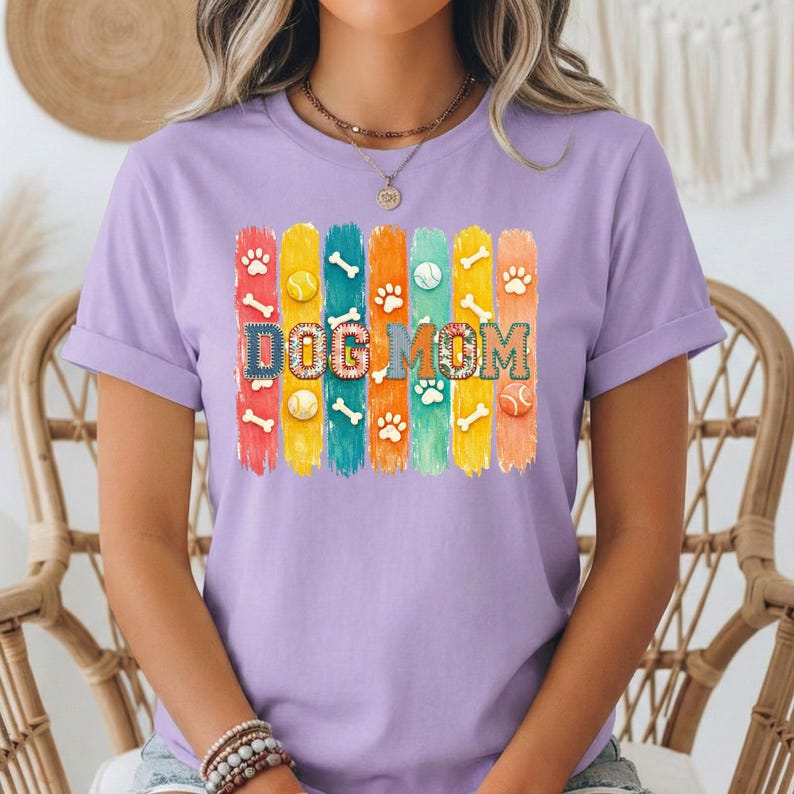 Colorful dog mom graphic with vertical painted stripes, paw prints, bones, and tennis balls in a playful patchwork style design for shirts and sublimation.