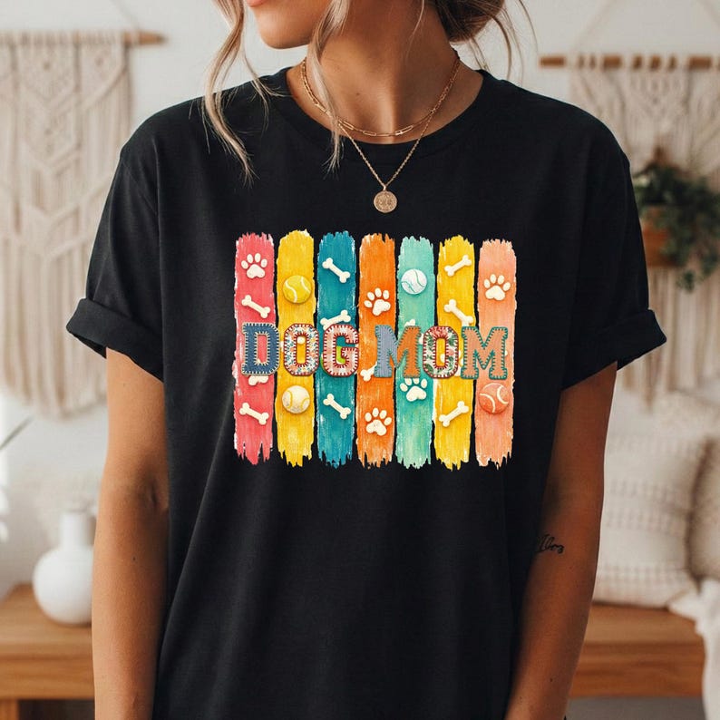 Colorful dog mom graphic with vertical painted stripes, paw prints, bones, and tennis balls in a playful patchwork style design for shirts and sublimation.