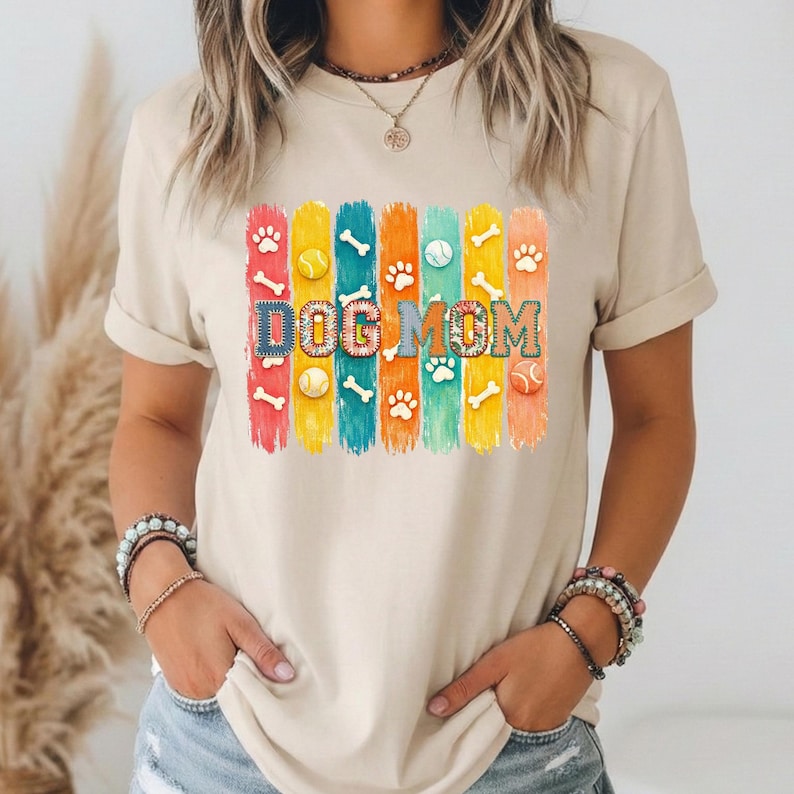 Colorful dog mom graphic with vertical painted stripes, paw prints, bones, and tennis balls in a playful patchwork style design for shirts and sublimation.